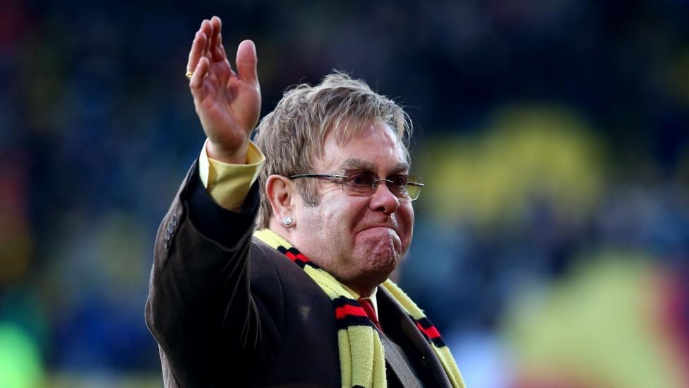 Elton John wearing a yellow scarf with red and black stripes, waving on the pitch at Vicarage Road