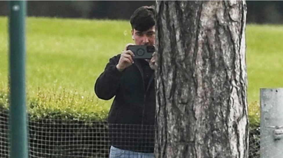 A man in a dark jacket and blue jeans stands partially concealed by a tree trunk and holds a phone up in two hands as if filming and has a white earphone in his right ear